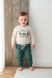 Child wearing a white sweater with text and green pants standing against a wooden wall.
