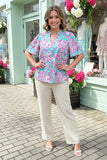 Woman wearing a colorful floral blouse and beige pants standing in front of a store with flowers.