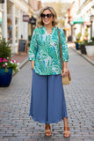 Woman in a green paisley blouse and blue pants walking on a street.