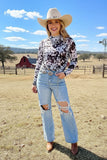 Woman in a cow print shirt, jeans, and cowboy hat standing in a rural landscape with a red barn and windmill.